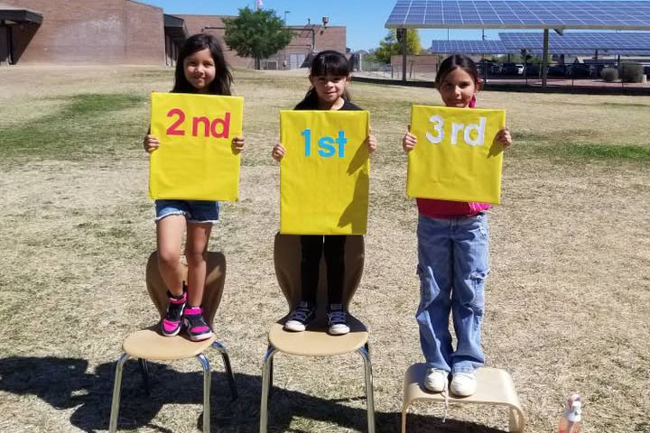 Three girls stand on chairs on the field holding signs that say 2nd, 1st and 3rd
