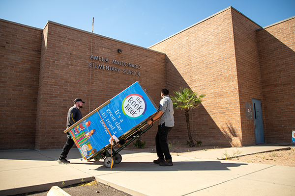 Two men wheel a book vending machine into the school