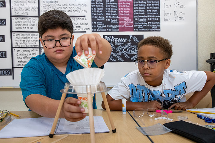 A boy with glasses pours something from a paper cup into a coffee filter, while another boy wearing glasses watches