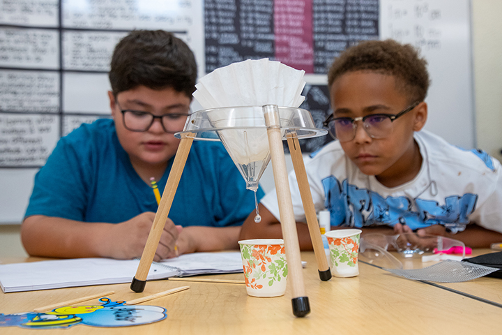 Two boys wearing glasses observe a liquid as it drips from a coffee filter into a paper cup