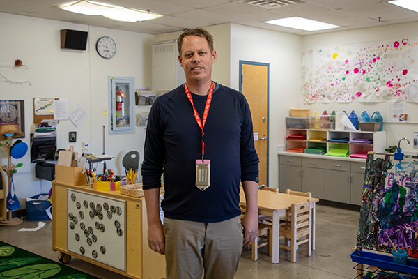 A man in a navy shirt and khakis smiles in his classroom