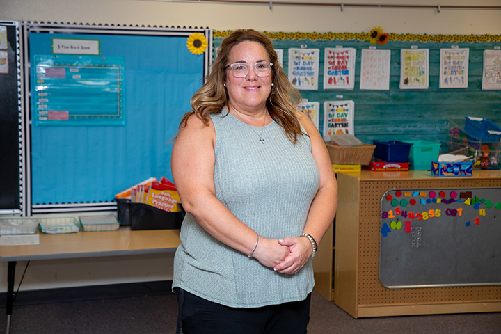 A woman with glasses and a gray tank top smiles in her classroom